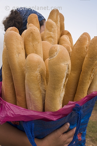 Woman in headscarf carries a shopping bag full of French baguette bread sticks.