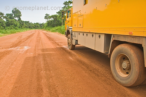 Yellow Oasis Overland truck parked by side of red dirt road in the jungle.