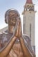 Image of Bronze painted statue of woman praying in front of the white stucco Roman Catholic church.