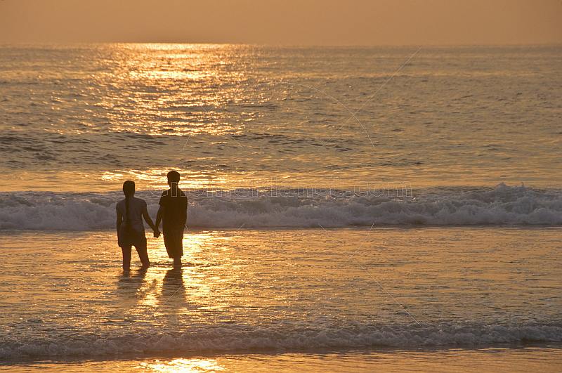 Young Indian couple hold hands in the surf at sunset.