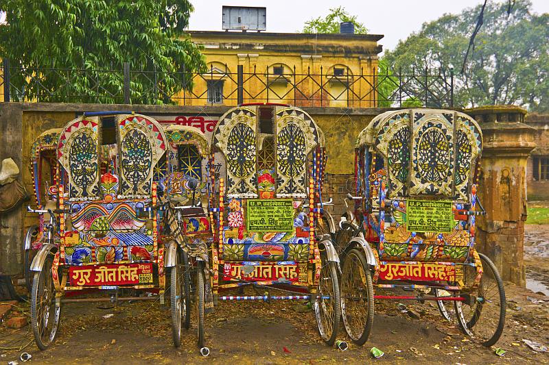 Three colorful decorated bicycle rickshaws next to old city wall.