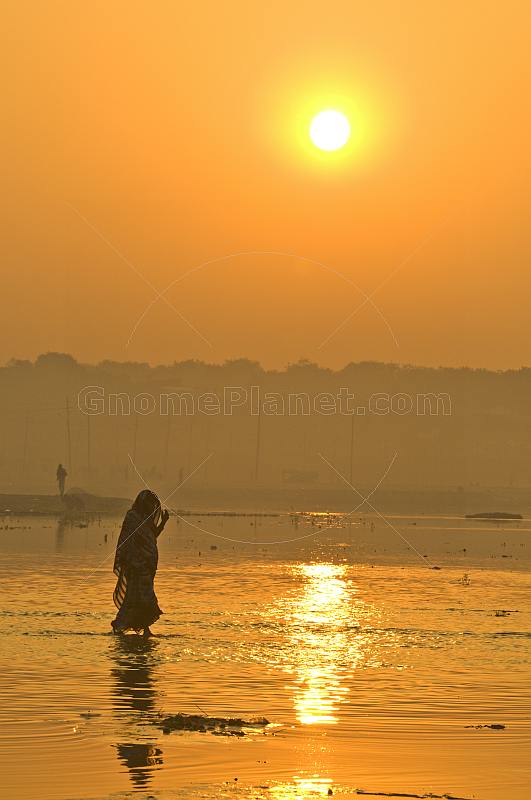 Woman in sari walks through Ganges River shallows at dawn for a ritual bath.