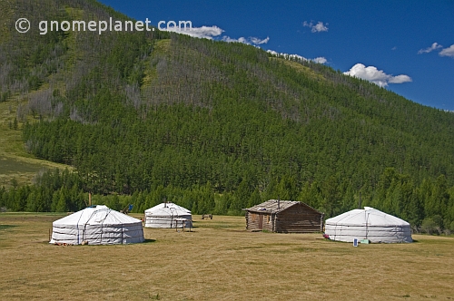 A group of yurts and a log cabin nestle in a forested mountain valley.