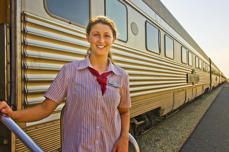 Smiling Great Southern Rail female attendant next to Ghan train carriages at Alice Springs station.