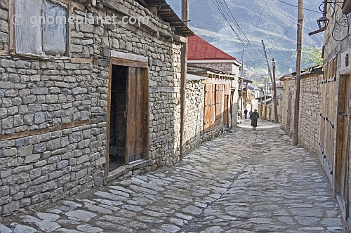 An old woman walks down the cobbled village street between houses protected from earthquake damage by wooden beams integrated in the stonework.