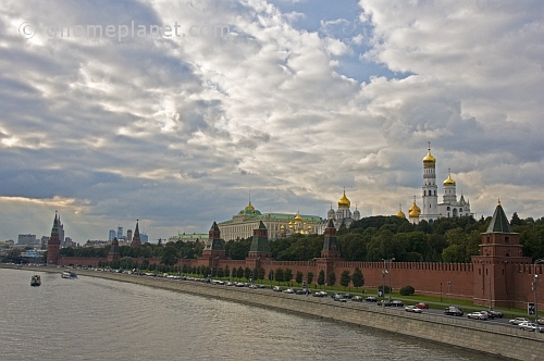 The golden domes and green copper roofs of the Kremlin along the Moscow River contrast a cloudy sky.