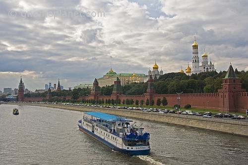 A sight-seeing boat on the Moscow River passes the red walls of the Kremlin, under a cloudy sky.