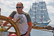 Man at the wheel of the 3 masted barque 'Picton Castle' with tallship 'Sagres' in background.