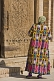 Elderly Uzbek lady in traditional hand dyed silk Chopon coat, stands before an intricately carved wooden door.