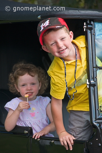 Small girl with lollipop and boy in yellow shirt wait in the back seat of a vintage car.