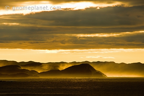 The early morning sun shines through clouds to light islands and ocean spray.