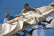 Two male crew members of the tallship 'Picton Castle' work aloft to stow sail.
