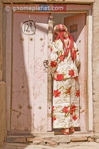 Uighur woman in white and red skirt and top, standing in pink doorway.
