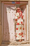Uighur woman in white and red skirt and top, standing in pink doorway.