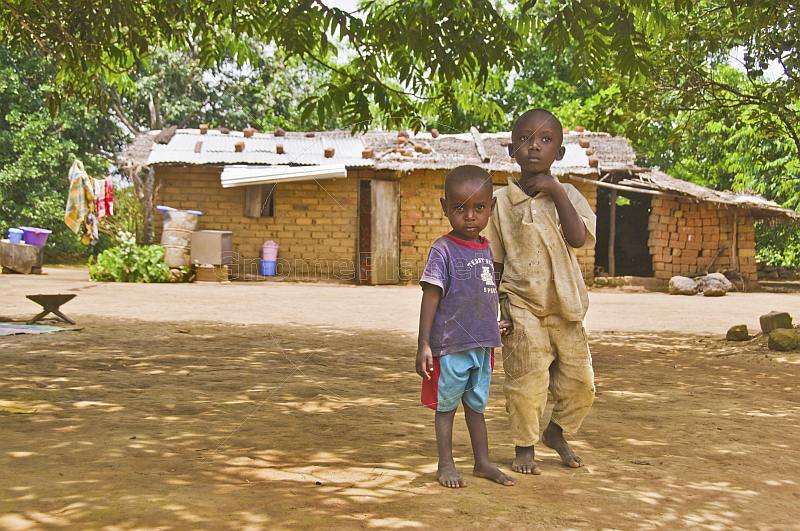 Two small brothers stand in front of their mud brick house.