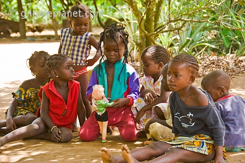 Group of children sit in the shade and play with a plastic doll.