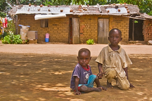 Two small brothers kneel in front of their mud brick house.