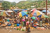 Women under umbrellas selling vegetables in a crowded market with views of hillside to rear.