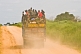 Image of A heavily laden truck with passengers riding on top of the cargo drives down a dusty main road.